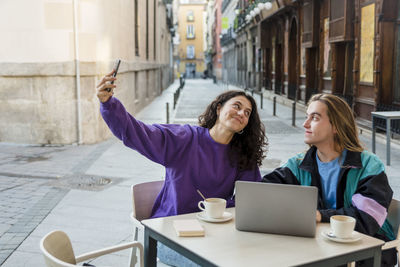 Friends taking selfies with a mobile phone while sitting at an outdoor cafe.