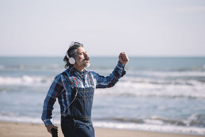 Portrait of happy man in jumpsuit dancing with headphones on the beach