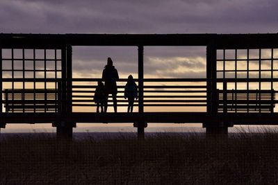 Silhouette people on railing against sky during sunset