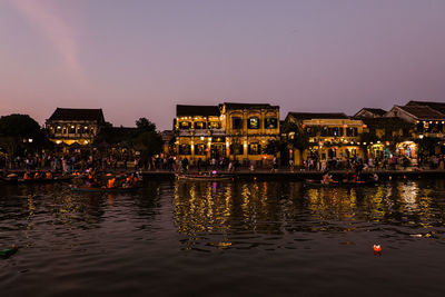 Illuminated buildings by lake against sky at dusk