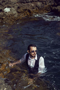 Man with a beard and sunglasses in clothes a vest and a white shirt swims in the sea among the rocks