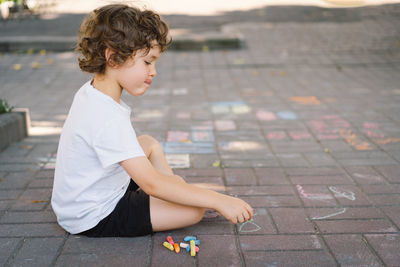 Little preschool boy draws with colorful chalks on the ground.