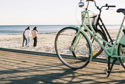 Full length of friends walking on beach with bicycles in foreground