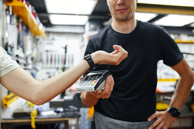 Soft focus of male mechanic with terminal receiving contactless payment from crop customer with smart watch during work in garage