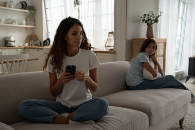 Caring puzzled mother sits on couch with phone and looks back at offended daughter after punishment