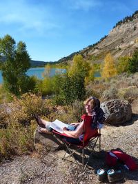 Woman resting while reading book by mountain against sky during sunny day