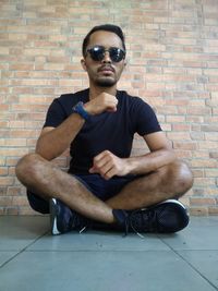 Young man wearing sunglasses sitting against brick wall