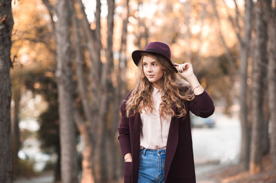 Woman wearing hat standing with hand in pockets at forest