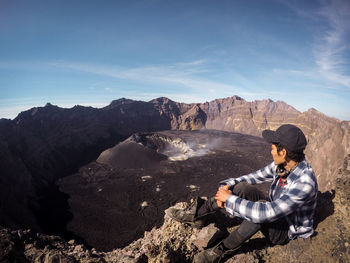 Man sitting on rock looking at mountains against sky