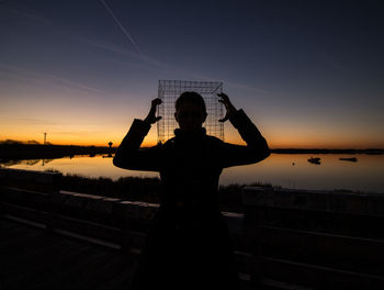 Silhouette man wearing metallic cage while standing on shore against sky during sunset