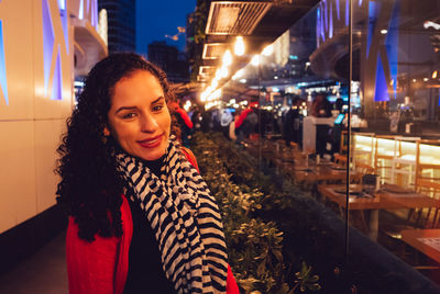 Portrait of smiling young woman by illuminated restaurant at night