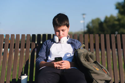 A cute boy teenager in headphones , watches mobile phone and sits on bench outdoor in the park 
