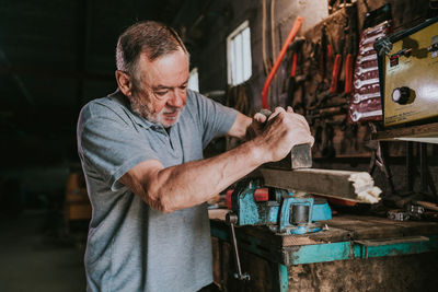 Focused mature male woodworker using jointer for planning wooden plank while working at shabby workbench in joinery workshop