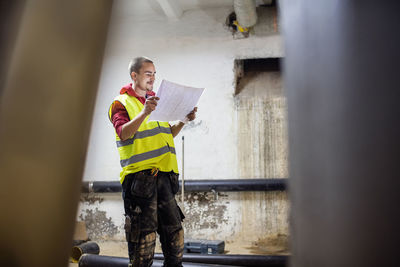 Smiling plumber reading document while standing by pipes in basement