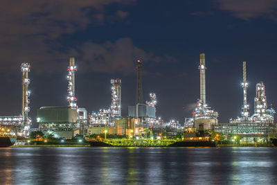 Illuminated factory by river against sky at night