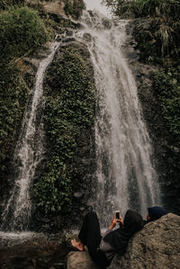 Couple lying down on rock while using mobile phone against waterfall
