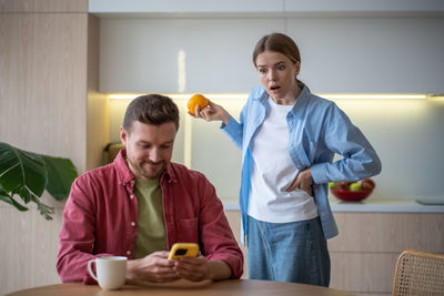 Amazed jealous shocked woman stands behind back peeping in man husband smartphone on kitchen at home