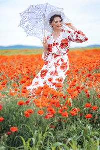 Happy woman in summer dress with a umbrella dances in the field with blooming poppies against a sky