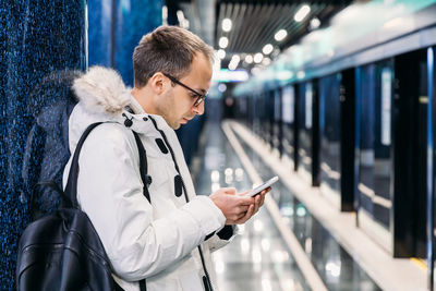 Handsome adult man in white jacket with glasses waiting subway train. killing time in mobile phone