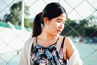 Young woman looking away standing against chainlink fence