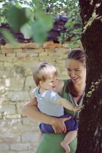 Happy cute blonde baby boy holded in arms by cute smiling women near tree