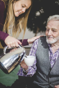 Young woman pouring drink in cup being held by grandfather in nursing home