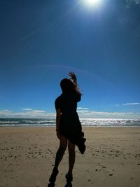 Silhouette woman with hand raised standing at beach against sky during sunny day