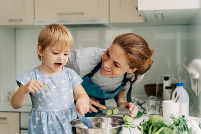 Funny toddler daughter helps mom cook in the home kitchen and eats vegetables.