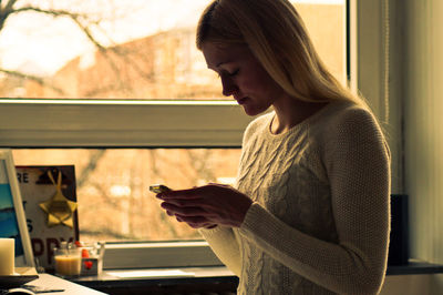 Woman using mobile phone against window at home