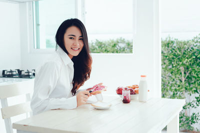 Portrait of happy woman applying preserves on bread slice at table during morning