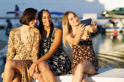 Female friends taking selfie through smart phone sitting on retaining wall