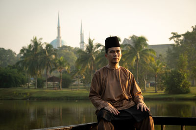 Portrait of man wearing traditional clothing while sitting on railing against lake during sunset