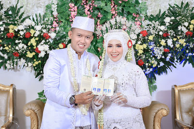 Portrait of a smiling young woman and man holding flowers, at their wedding