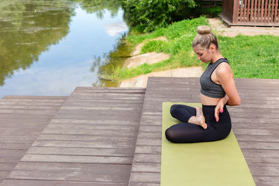 A slender woman sitting on a wooden platform by a pond in the park in summer and performing yoga