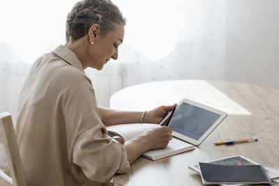 Woman holding digital tablet with data and taking notes at table at home