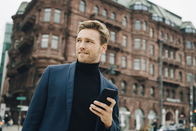 Confident businessman with smart phone looking away while standing against building in city