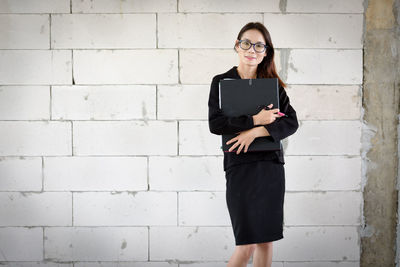 Portrait of young businesswoman with file standing against wall at construction site