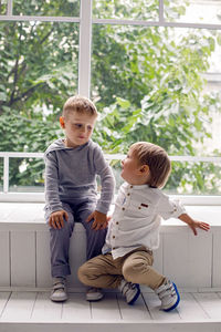 Two brothers sitting by the window in a white studio with green trees on the street