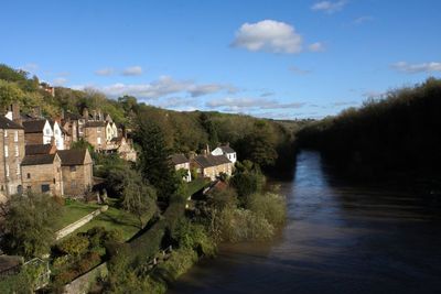 Houses by river amidst trees and buildings against sky