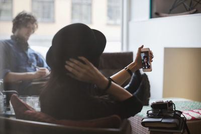 Rear view of woman taking selfie with colleague working in background seen through glass