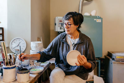 Skilled middle aged female artist coloring handmade clay bowl with paintbrush and paints while working in workshop