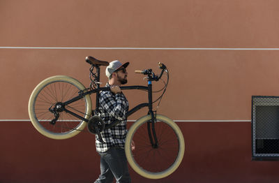 Side view of man carrying bicycle while walking against building in city during sunny day