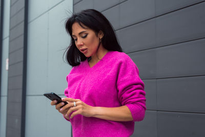 Beautiful woman wearing pink sweater posing on dark gray background at city street with smartphone