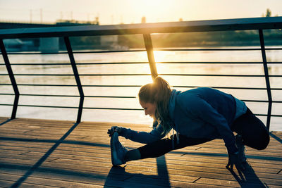 Young woman exercising on promenade by river against clear sky during sunset