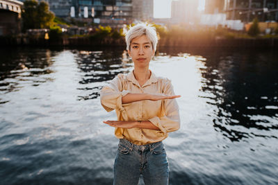 Portrait of young woman gesturing equal sign while standing against lake in city