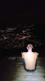 Rear view of shirtless man sitting in swimming pool at night