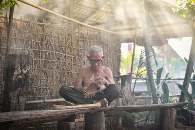 Shirtless man weaving wicker in hut