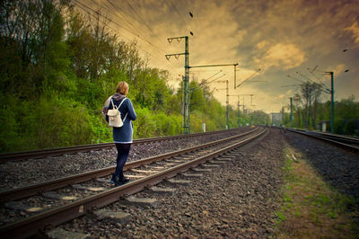 Rear view of backpack woman walking on railroad tracks against cloudy sky