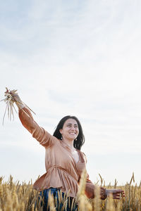 Mid adult woman in beige shirt walking across golden field holding heap of rye lit by sunset light