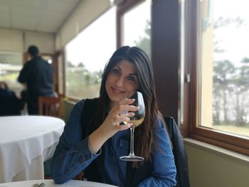 Portrait of young woman holding wineglass while sitting by window in restaurant